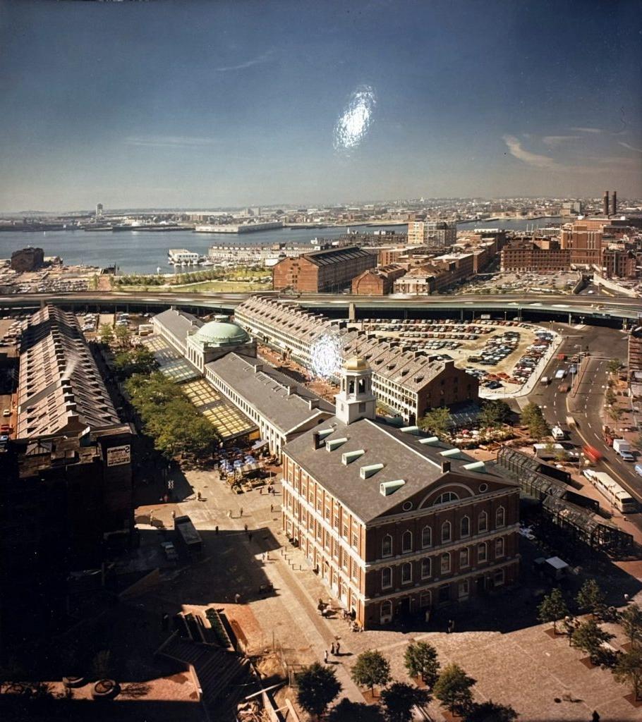 An aerial view of Faneuil Hall and Quincy Market. Pedestrians walk around the buildings, while roads and highways stretch to the sides and rear of the marketplace. Boston Harbor is visible in the background.